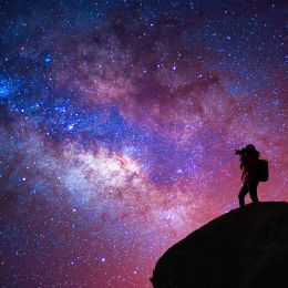 person standing on a cliff photographing a colorful night sky galaxy
